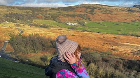 Alex is standing sideways on a mountain wearing a wooly hat. She is pulling her hat down with her hands and there is blue sky and hills in the background.